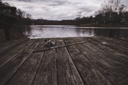 Téléchargez les photos : Une vieille canne à pêche abandonnée repose sur un quai en bois avant le lac - en image libre de droit