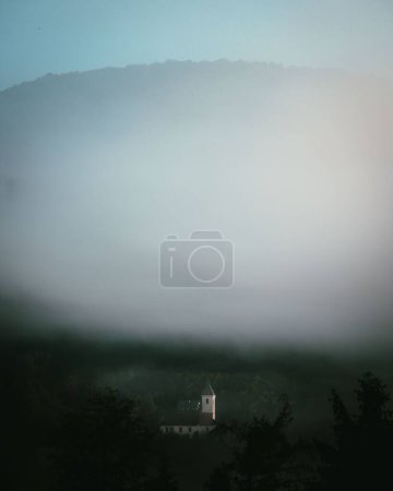 Téléchargez les photos : Une grande masse de brouillard dans le ciel rassemblée au-dessus d'une tour de bâtiment dans un paysage verdoyant - en image libre de droit