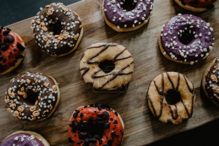Foto de Una vista superior de donuts recién horneados, glaseados con chocolate y vainilla, decorados de manera diferente, servidos en una tabla de cortar de madera - Imagen libre de derechos