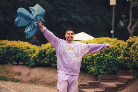 Téléchargez les photos : Un homme hispanique heureux dans un costume de sport violet tie-dye marchant dans un parc avec des ballons bleus - en image libre de droit