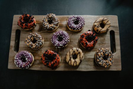 Foto de Una vista superior de donuts recién horneados, glaseados con chocolate y vainilla, decorados de manera diferente, servidos en una tabla de cortar de madera - Imagen libre de derechos