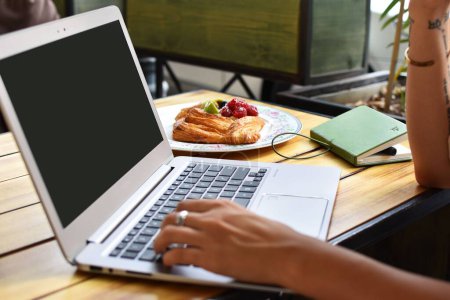 Téléchargez les photos : Gros plan d'une jeune femme travaillant sur son ordinateur portable et ayant un croissant dans un café - en image libre de droit