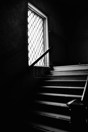 Photo for A vertical grayscale of stairs and a window with railings in a dark building - Royalty Free Image