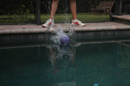 Téléchargez les photos : Une balle éclaboussant dans l'eau de la piscine sur le fond des jambes d'une personne debout au bord de la piscine - en image libre de droit