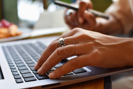 Téléchargez les photos : Gros plan d'une jeune femme travaillant sur son ordinateur portable et ayant un croissant dans un café - en image libre de droit