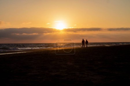 Photo for A silhouette view of a couple walking on the beach by the sea under orange sunset scene - Royalty Free Image