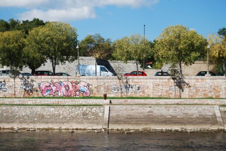 Foto de Una cerca de piedra con grafitti por el río Nisava y coches en la calle con árboles bajo el cielo azul - Imagen libre de derechos