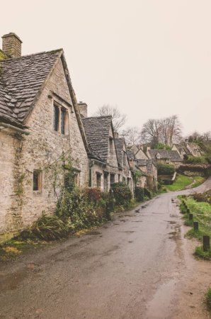 Photo for A vertical shot of a wet road running alongside a row of historic quintessential Cotswold cottages in Bibury, England - Royalty Free Image