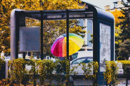 Foto de Una persona con paraguas de arco iris esperando en la estación de autobuses en otoño - Imagen libre de derechos