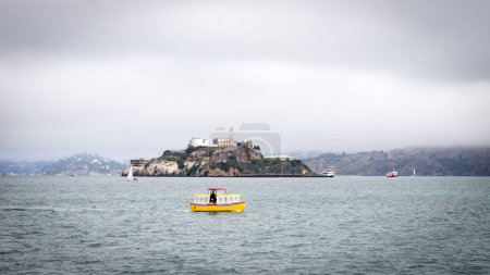Foto de Alcatraz en una mañana nublada con una pequeña embarcación amarilla pasando por el centro de primer plano. - Imagen libre de derechos