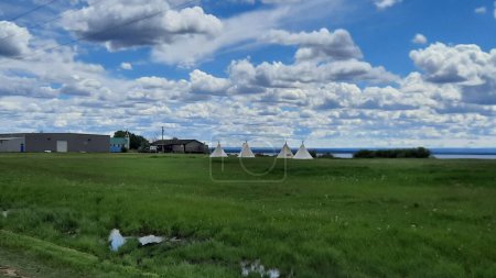 Foto de Una hermosa toma de tipis montada en verdes llanuras, bajo un cielo azul claro con nubes blancas dispersas - Imagen libre de derechos