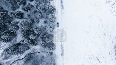 Téléchargez les photos : Un drone d'une voiture conduisant sur une route recouverte de neige à côté d'une forêt d'épinettes - en image libre de droit