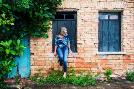 Photo for Woman leaning against a window of an old house. Brick facade and leaves around. Aratuipe, Bahia. - Royalty Free Image