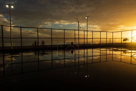 Foto de Silueta de personas ejercitándose en el borde de la playa de Río Vermelho en Salvador, Bahía, contra la puesta de sol amarilla. - Imagen libre de derechos