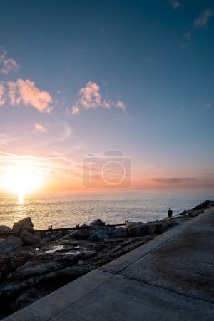 Téléchargez les photos : Un plan vertical de la belle scène de la nature de la plage rocheuse et une personne debout et regardant la mer au coucher du soleil - en image libre de droit
