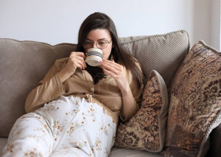 Photo for A beautiful pregnant Caucasian woman drinking tea while sitting on a couch - Royalty Free Image