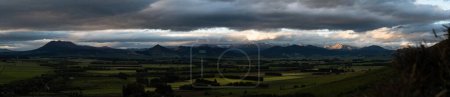 Téléchargez les photos : Vue panoramique d'un ciel nuageux sombre avec le soleil qui brille sur les champs verts ruraux - en image libre de droit