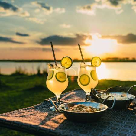 Téléchargez les photos : Un gros plan de cocktails au citron avec des apéritifs sur un fond de lac lors d'un coucher de soleil - en image libre de droit
