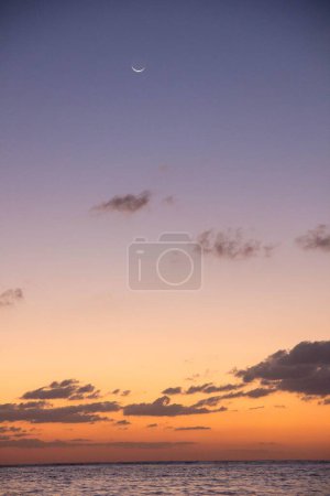 Foto de Una hermosa vista de un océano durante una puesta de sol con nubes de ensueño y una creciente luna creciente en el cielo - Imagen libre de derechos
