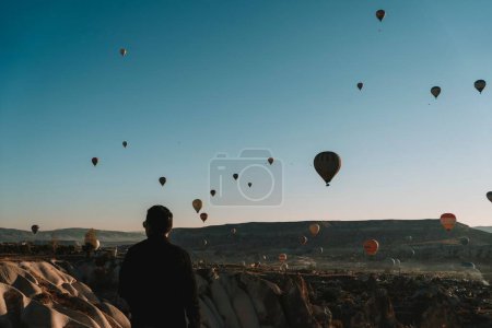 Téléchargez les photos : Un homme regarde voler un ballon à air chaud dans le ciel bleu en Cappadoce, Turquie - en image libre de droit