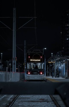 Foto de Una foto vertical de la calle de un tranvía en una estación con gente alrededor en Kitchener que conduce a Conestoga por la noche - Imagen libre de derechos