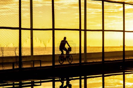 Foto de Salvador, Bahia, Brasil - 05 de diciembre de 2021: Ciclista haciendo ejercicio al borde de la playa de Río Vermelho contra la puesta de sol. Ciudad de Salvador, Bahia. - Imagen libre de derechos