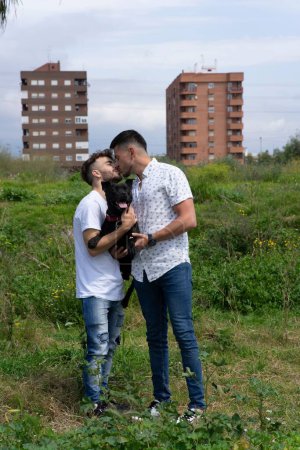 Foto de Una toma vertical de una pareja gay hispana besándose con su perro al aire libre. - Imagen libre de derechos