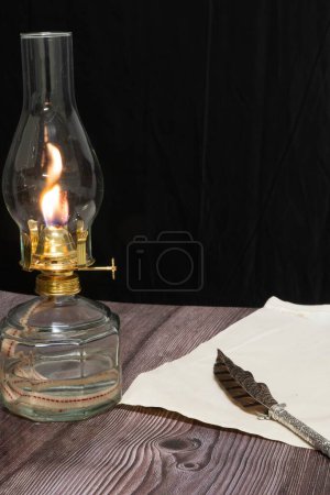 Foto de Primer plano vertical de una vieja lámpara de aceite de huracán antigua en llamas con pluma de pluma y papel sobre una mesa de madera - Imagen libre de derechos