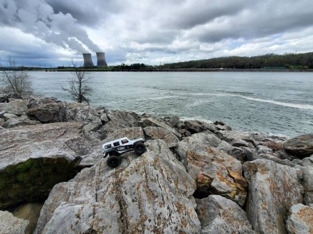 Téléchargez les photos : Une voiture jouet Jeep Wrangler sur des pierres au bord du lac avec vue sur la centrale nucléaire - en image libre de droit
