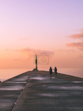 Téléchargez les photos : Une belle scène de nature avec un trottoir sur la plage avec des gens qui marchent et un phare au coucher du soleil - en image libre de droit