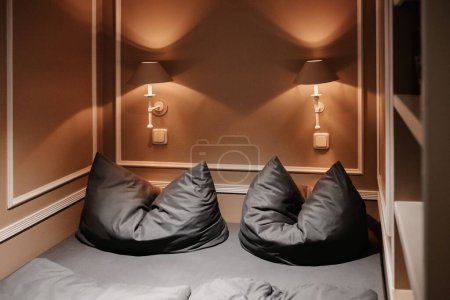 Téléchargez les photos : Un lit avec deux oreillers gris et des lampes sur les murs bruns dans une petite pièce - en image libre de droit