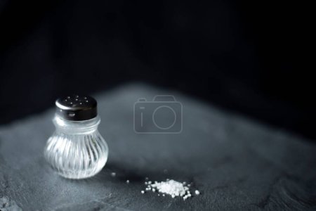 Foto de Un primer plano de un salero de vidrio y algunos granos de sal esparcidos sobre una superficie gris - Imagen libre de derechos