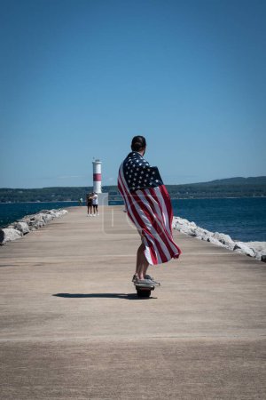 Photo for A closeup view of a man skateboarding with an American flag on his shoulder along the stone pier - Royalty Free Image