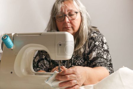 Photo for Older woman with white hair out of focus sewing a white fabric on a sewing machine - Royalty Free Image