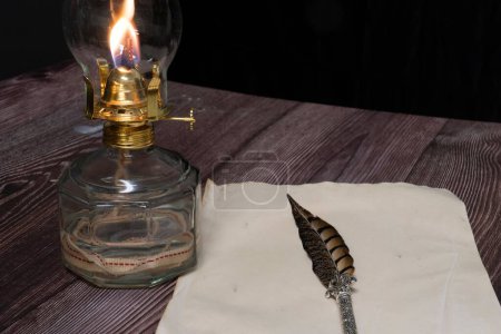 Foto de Un primer plano de una vieja lámpara de aceite de huracán antigua en llamas con pluma de pluma y papel sobre una mesa de madera - Imagen libre de derechos