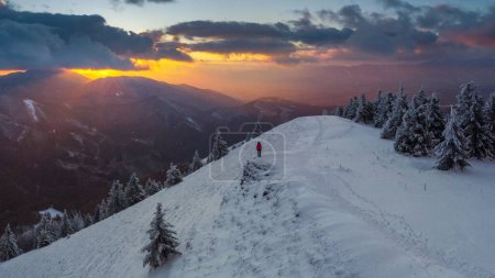 Foto de Un pintoresco plano de un excursionista caminando en la cima de una montaña nevada a lo largo de abetos al atardecer - Imagen libre de derechos