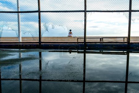 Foto de Una persona sentada en la balaustrada frente al mar de la playa de Río Vermelho en la ciudad de Salvador, Bahía, durante la tarde. - Imagen libre de derechos