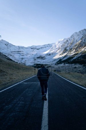 Téléchargez les photos : Plan vertical d'un homme marchant le long d'une route asphaltée contre les collines enneigées - en image libre de droit