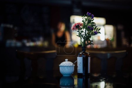 Foto de Florero con flores en la mesa de la cafetería junto a la olla de azúcar con persona borrosa en el fondo y la iluminación de mal humor en la barra hipster - Imagen libre de derechos