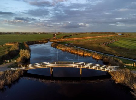 Téléchargez les photos : Un pont incurvé au-dessus d'une rivière avec son image reflétant dans l'eau, dans une prairie verte - en image libre de droit