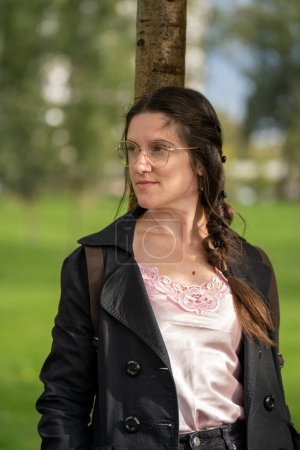 Photo for A vertical shot of a white female wearing specs and posing for the camera leaning on a tree - Royalty Free Image