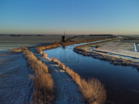 Téléchargez les photos : Un paysage hivernal d'une rivière le long des champs gelés et un vieux moulin à vent en arrière-plan - en image libre de droit