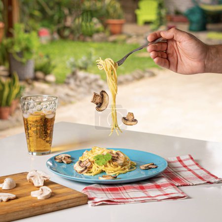 Photo for A man holding a fork with spaghetti and falling mushrooms above the plate next to a glass of juice and a wooden board with sliced mushrooms - Royalty Free Image