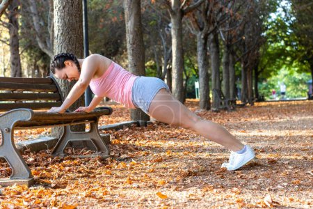 Téléchargez les photos : Une femme en tenue de sport faisant des pompes sur un banc de parc un jour d'automne calme - en image libre de droit