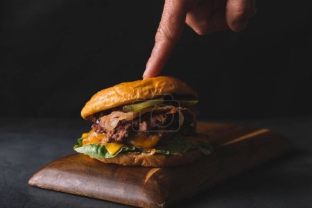 Photo for A close-up of a man touching a hamburger on a wooden board on a black background - Royalty Free Image