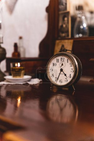 Photo for A vertical closeup of an old clock on a wooden table in an old house, old things blurred in the background - Royalty Free Image