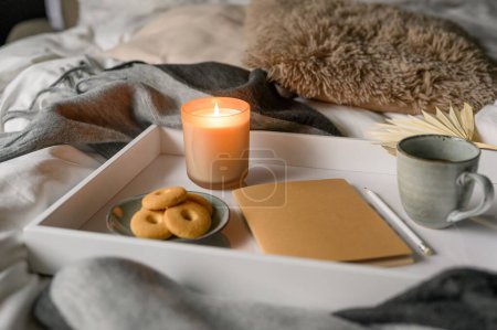 Foto de Acogedora composición de otoño con una taza de café, vela, galletas, cuaderno, lápiz en la cama cómoda - Imagen libre de derechos