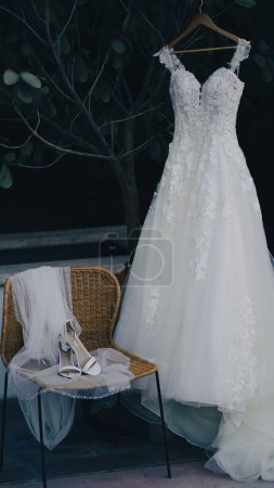 Foto de Un disparo vertical de un vestido de novia blanco en una percha cerca de los zapatos y un velo en una silla - Imagen libre de derechos