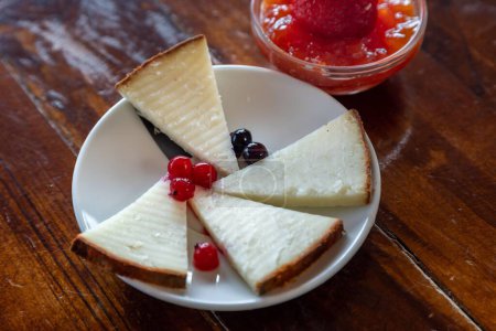 Foto de Un primer plano horizontal de cuatro piezas de pastel de queso en un plato blanco y mermelada de fresa en una mesa de madera - Imagen libre de derechos