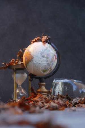 Photo for A low-angle shot of a globe and an hourglass on a table covered by autumn leaves, with a black background, on a sunny day outside - Royalty Free Image